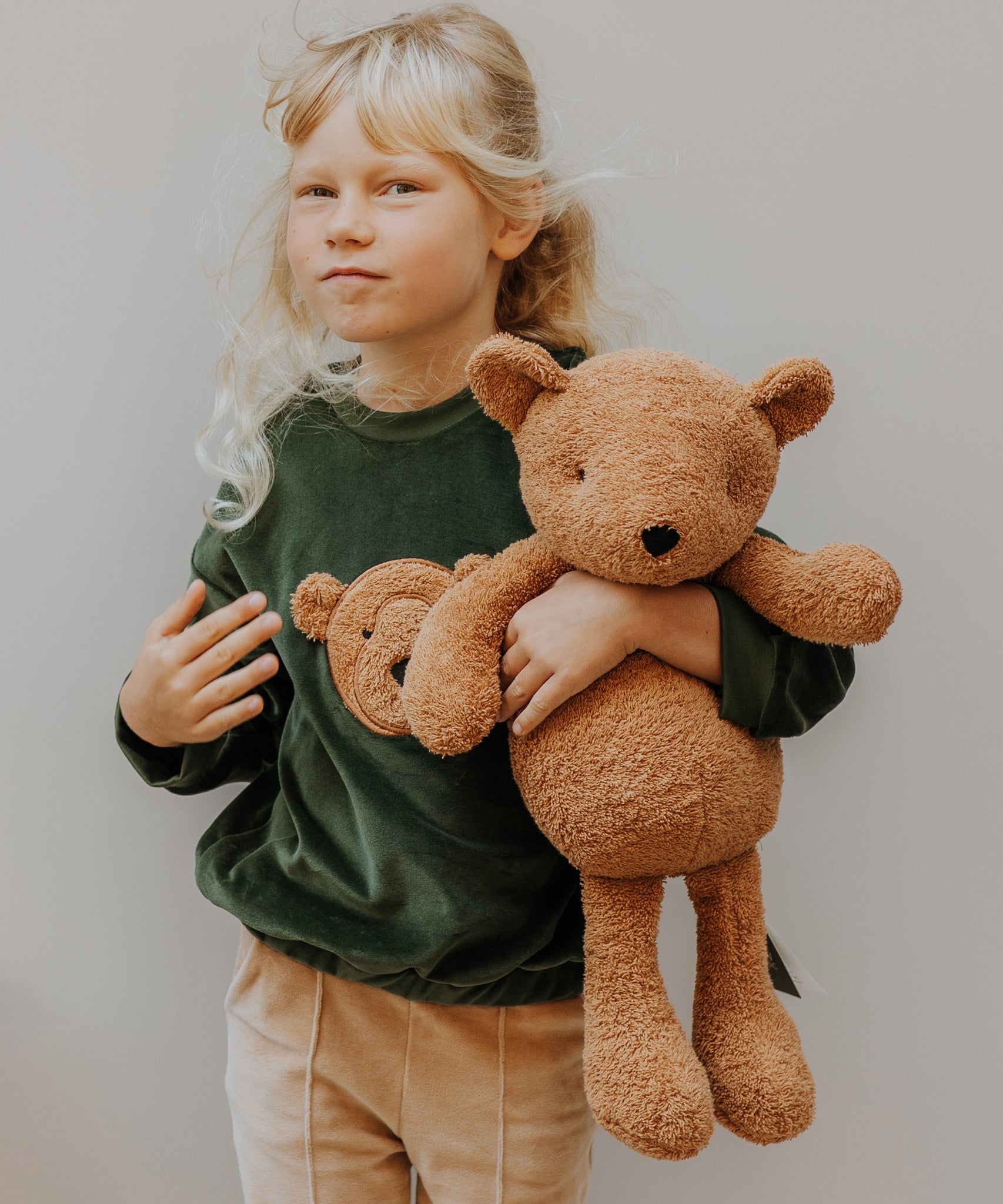 Child holding a large Wooly Organic teddy bear toy, made from organic cotton, in a caramel brown, for sale at Babipur.
