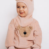 A close up of a child sitting down and wearing the Wooly Organic GOTS organic cotton embroidered velour sweater in pale blush pink /savannah available at Babipur. Showing the cute fluffy teddy bear embroidery on the front