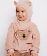 A close up of a child sitting down and wearing the Wooly Organic GOTS organic cotton embroidered velour sweater in pale blush pink /savannah available at Babipur. Showing the cute fluffy teddy bear embroidery on the front