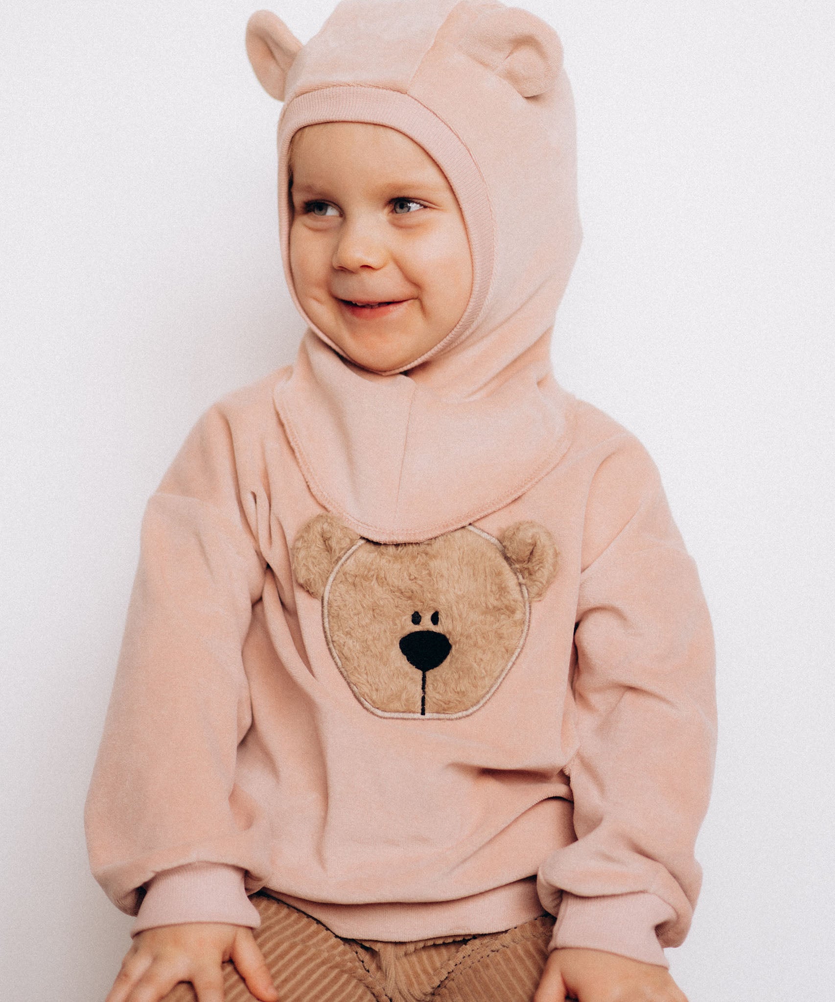 A close up of a child sitting down and wearing the Wooly Organic GOTS organic cotton embroidered velour sweater in pale blush pink /savannah available at Babipur. Showing the cute fluffy teddy bear embroidery on the front