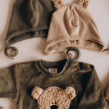 A flatlay showing the Wooly Organic hat with teddy bear ears in tea leaf /savannah next to the velour sweater, both  available at Babipur 
