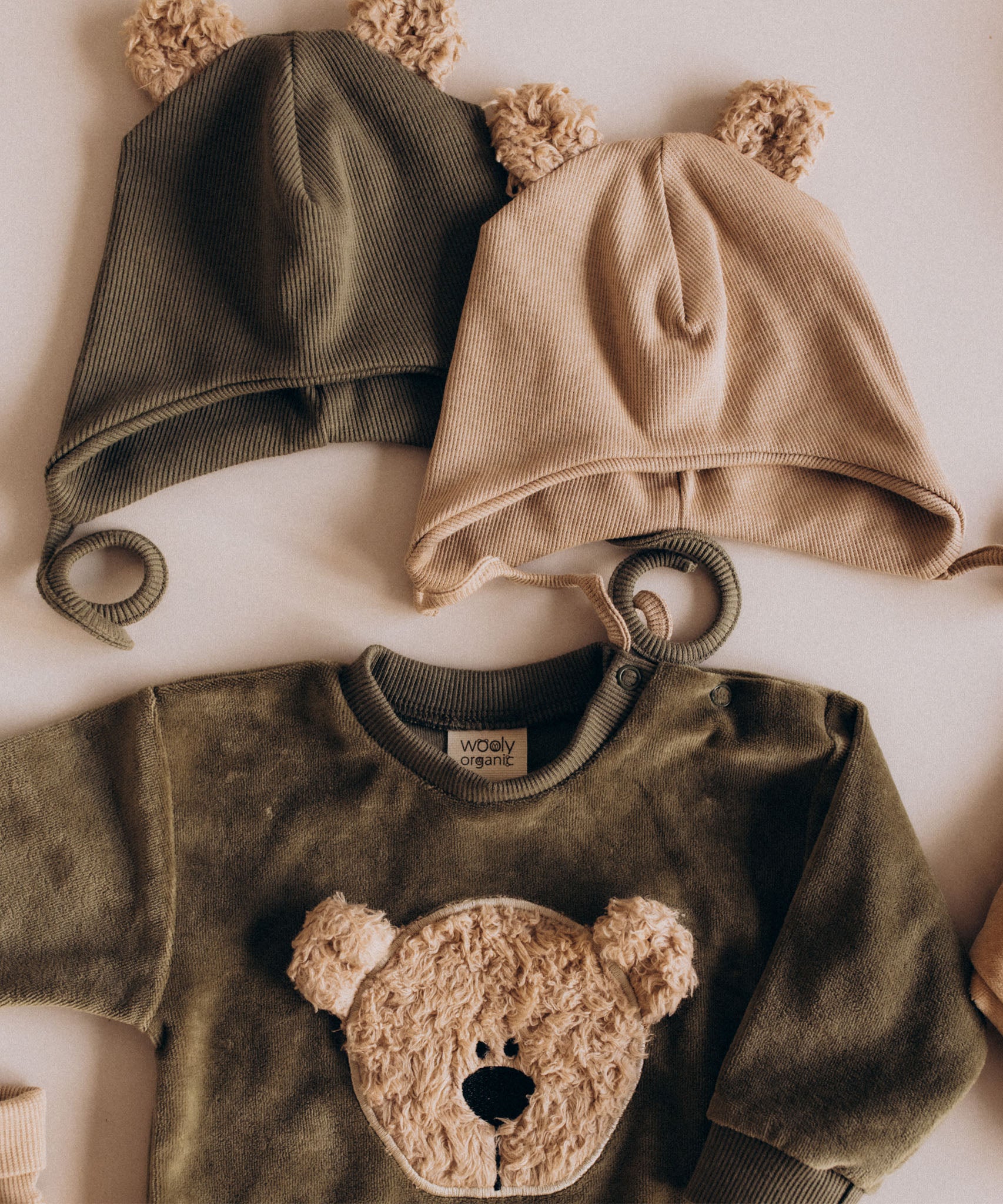 A flatlay showing the Wooly Organic hat with teddy bear ears in tea leaf /savannah next to the velour sweater, both  available at Babipur 