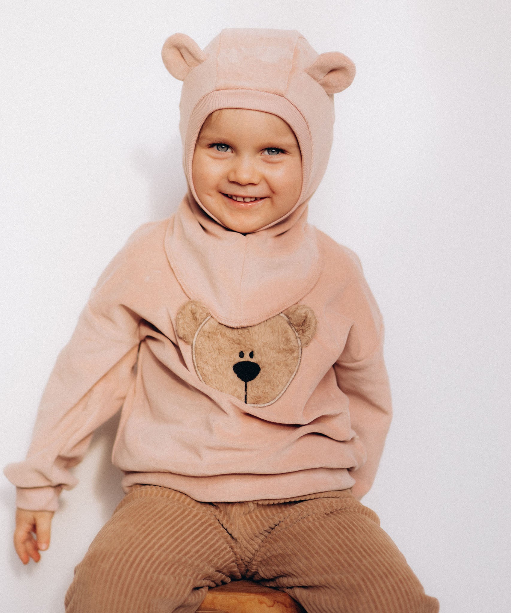 A child sitting down and wearing the Wooly Organic GOTS organic cotton embroidered velour sweater in pale blush pink /savannah available at Babipur