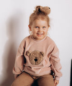 A child sitting down and wearing the Wooly Organic embroidered velour sweater in pale blush pink /savannah available at Babipur. 