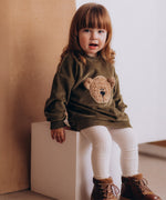 A child sitting on a wooden box and wearing the Wooly Organic embroidered GOTS organic cotton velour sweater in tea leaf /savannah available at Babipur. 