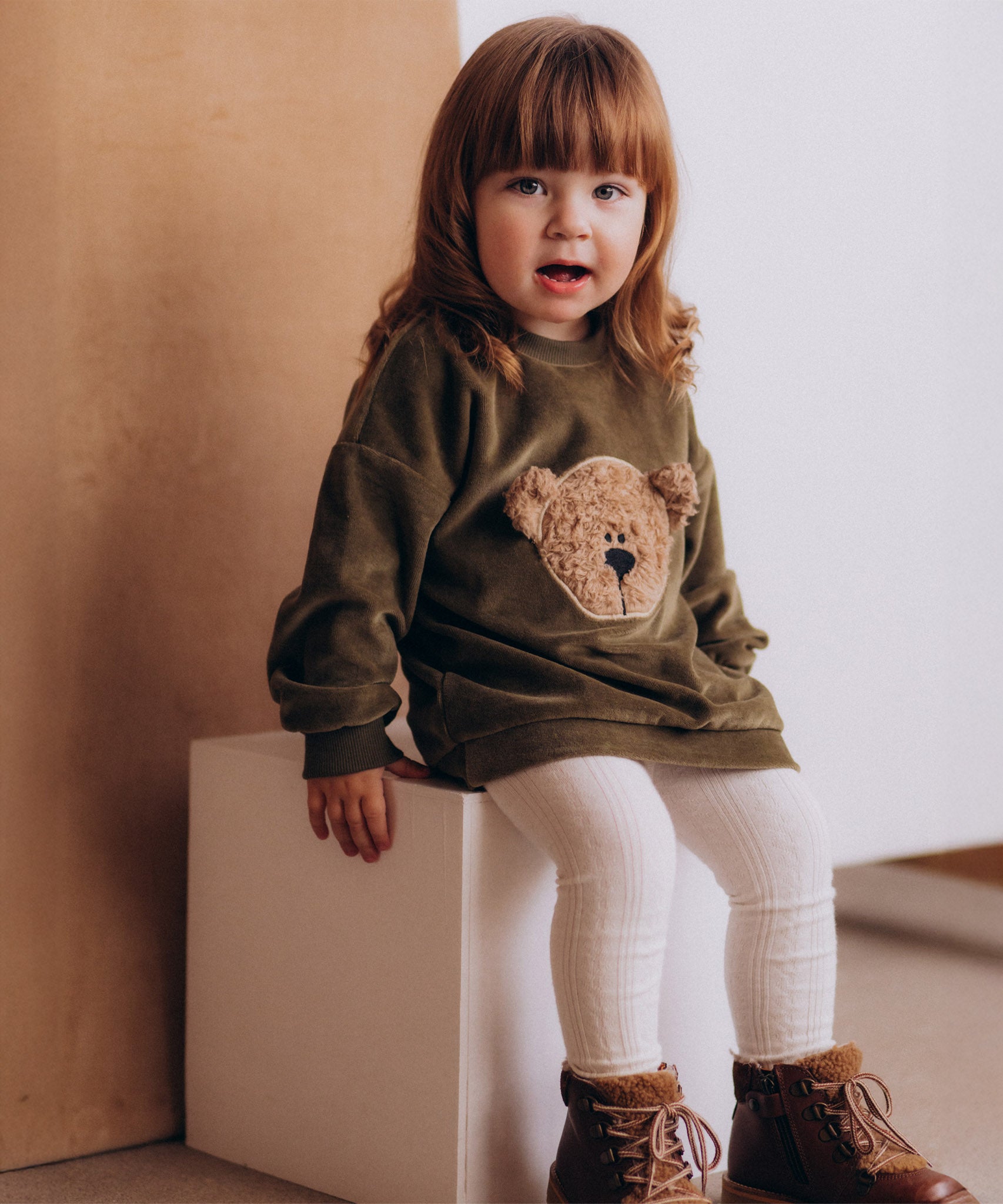 A child sitting on a wooden box and wearing the Wooly Organic embroidered GOTS organic cotton velour sweater in tea leaf /savannah available at Babipur. 