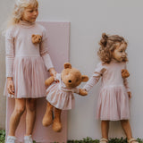 2 children standing on a grassy surface  and holding the Wooly Organic soft teddy bear in caramel brown with a pink tutu by the hands. Teddy bear available at Babipur. 