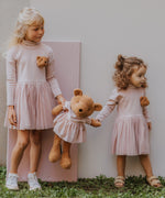 2 children standing on a grassy surface  and holding the Wooly Organic soft teddy bear in caramel brown with a pink tutu by the hands. Teddy bear available at Babipur. 