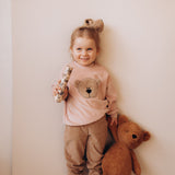 A child holding the Wooly Organic soft teddy bear and wearing the velour hair scrunchy, both available at Babipur.