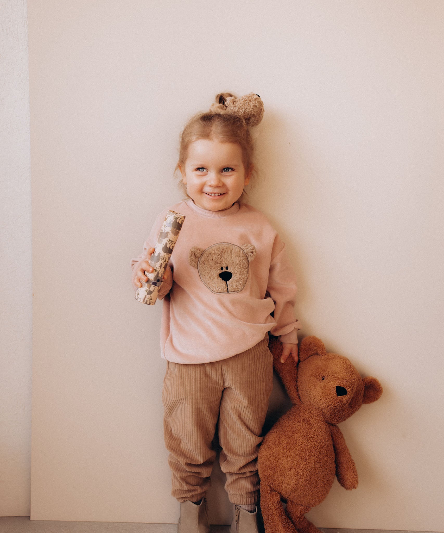 A child holding the Wooly Organic soft teddy bear and wearing the velour hair scrunchy, both available at Babipur.