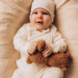 A baby lying on a wooly blanket and holding onto a small Wooly Organic soft teddy bear in brown available at Babipur. 