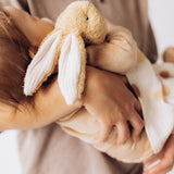  A mother holding a sleeping baby with a small Wooly Organic soft bunny rabbit toy in light brown available at Babipur. A soft cotton bunny rabbit with cute floppy ears