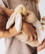  A mother holding a sleeping baby with a small Wooly Organic soft bunny rabbit toy in light brown available at Babipur. A soft cotton bunny rabbit with cute floppy ears