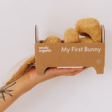 An adult hand holding the cardboard packaging shaped like a bed with the  A mother holding a sleeping baby with a small Wooly Organic soft bunny rabbit toy in light brown inside. Bunny rabbit available at Babipur. 