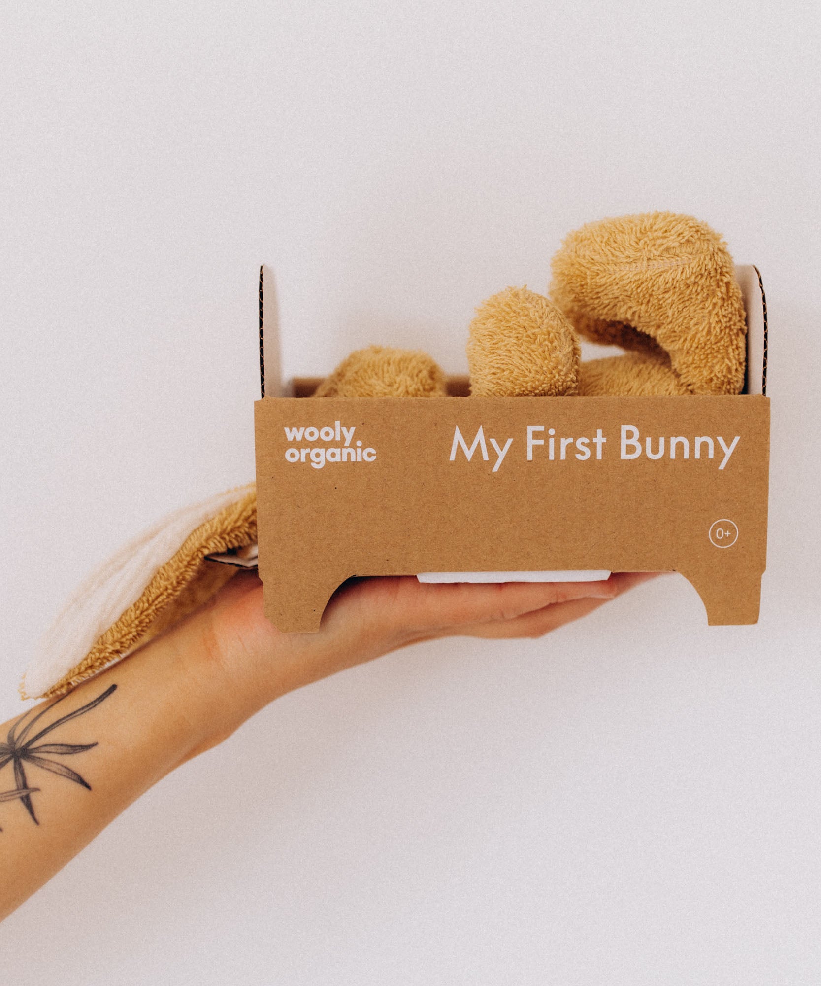 An adult hand holding the cardboard packaging shaped like a bed with the  A mother holding a sleeping baby with a small Wooly Organic soft bunny rabbit toy in light brown inside. Bunny rabbit available at Babipur. 