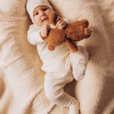A baby lying down on a soft blanket and playing with a small Wooly Organic soft teddy bear in brown available at Babipur. 