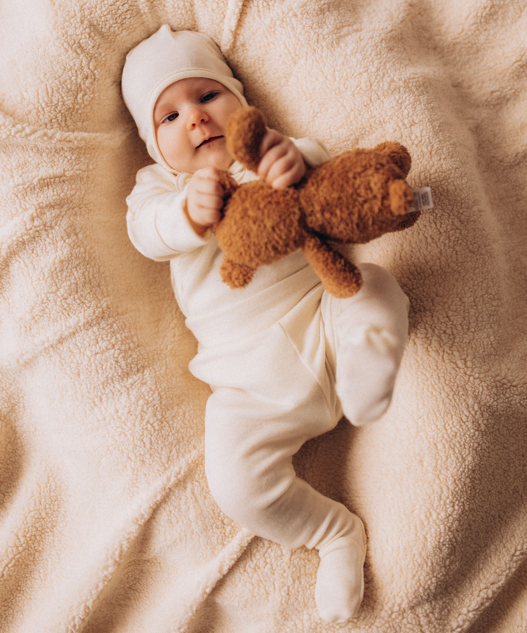 A baby lying down on a soft blanket and playing with a small Wooly Organic soft teddy bear in brown available at Babipur. 