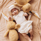  A baby lying on a blanket next to 2  Wooly Organic soft cotton bunny rabbit toys in light brown colour by the floppy ears. Bunny rabbit available at Babipur. 