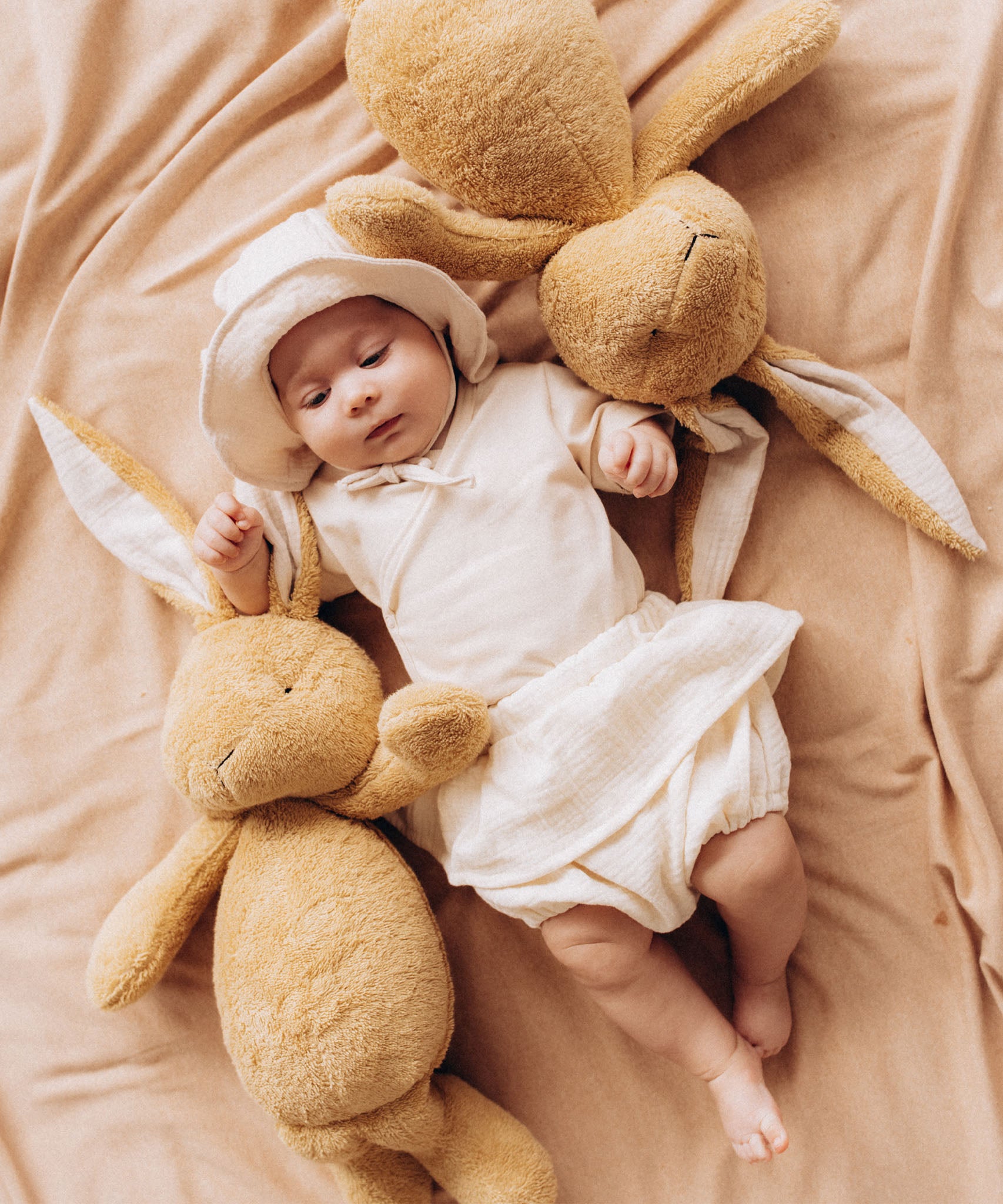  A baby lying on a blanket next to 2  Wooly Organic soft cotton bunny rabbit toys in light brown colour by the floppy ears. Bunny rabbit available at Babipur. 