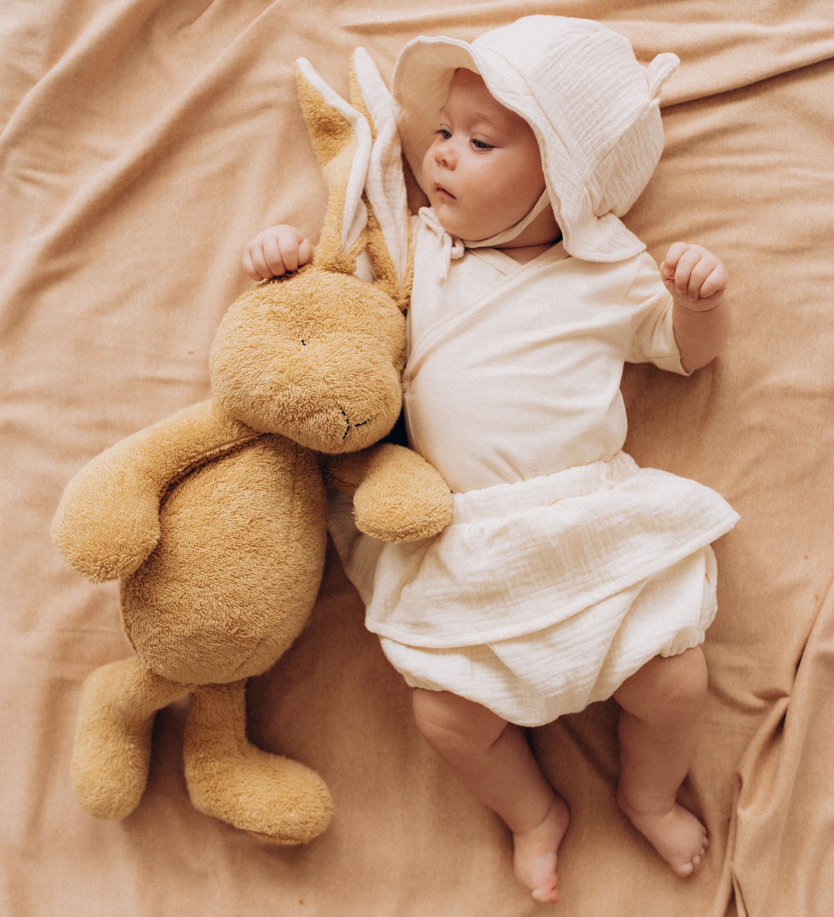 A baby lying next to a Wooly Organic soft cotton bunny rabbit toy in light brown colour available at Babipur. 