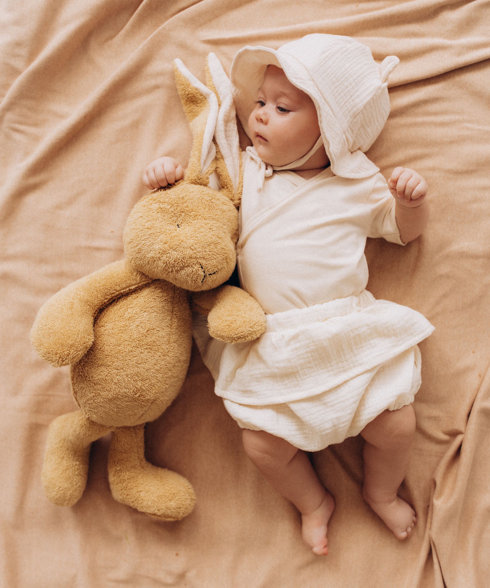 A baby lying next to a Wooly Organic soft cotton bunny rabbit toy in light brown colour available at Babipur. 