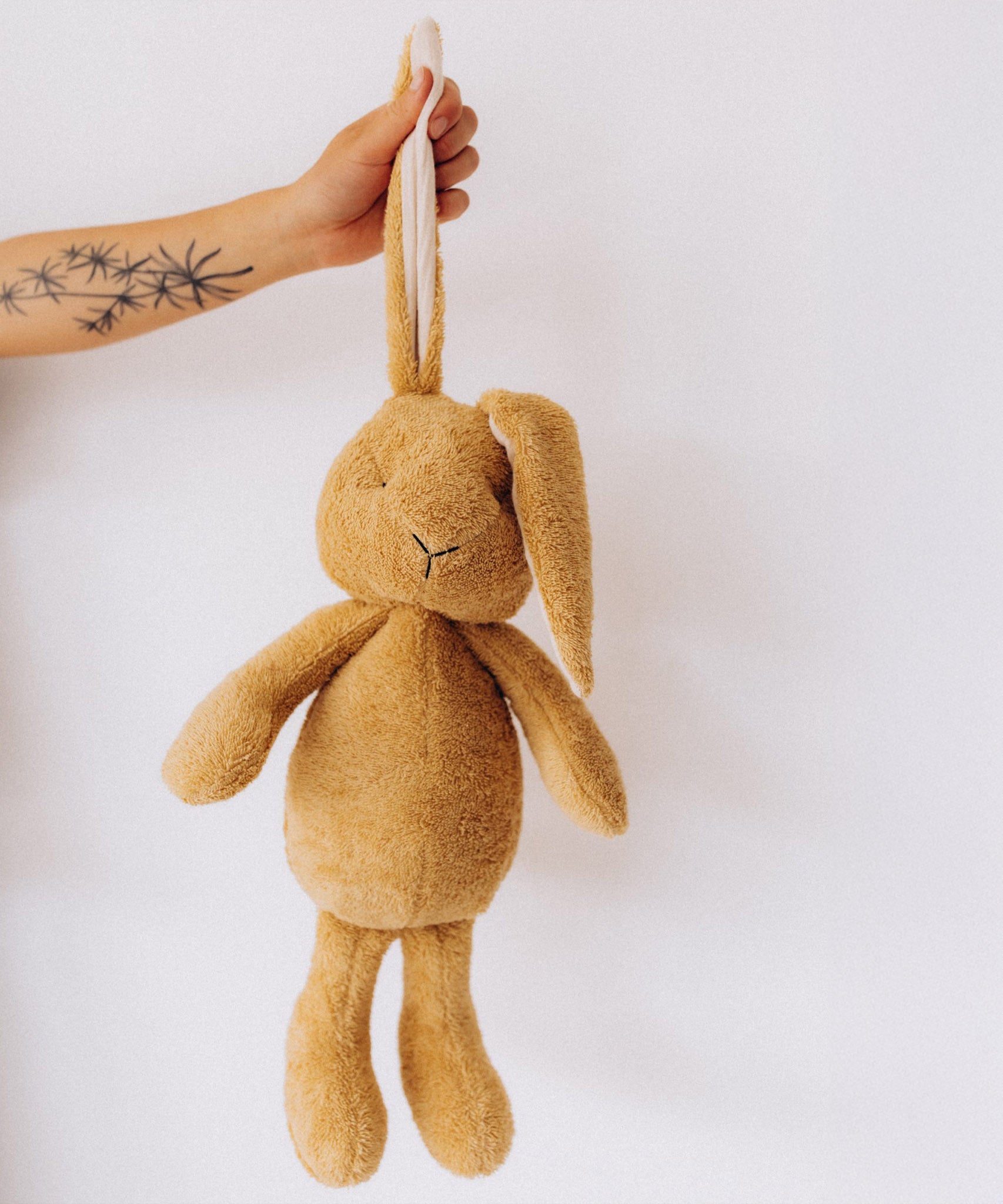 A adult holding the Wooly Organic soft cotton bunny rabbit toy in light brown colour by the floppy ears. Bunny rabbit available at Babipur. 