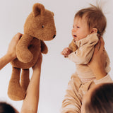 A baby being held next to a Wooly Organic soft teddy bear in caramel brown available at Babipur.