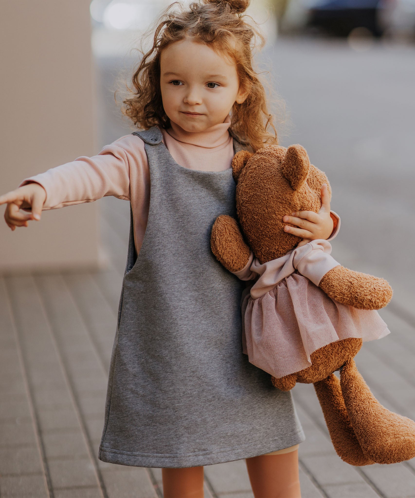 A child walking down a street holding the Wooly Organic soft teddy bear in caramel brown with a pink tutu available at Babipur. 