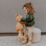 A child standing on a stone surface and playing with the Wooly Organic soft teddy bear in caramel available at Babipur