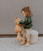 A child standing on a stone surface and playing with the Wooly Organic soft teddy bear in caramel available at Babipur