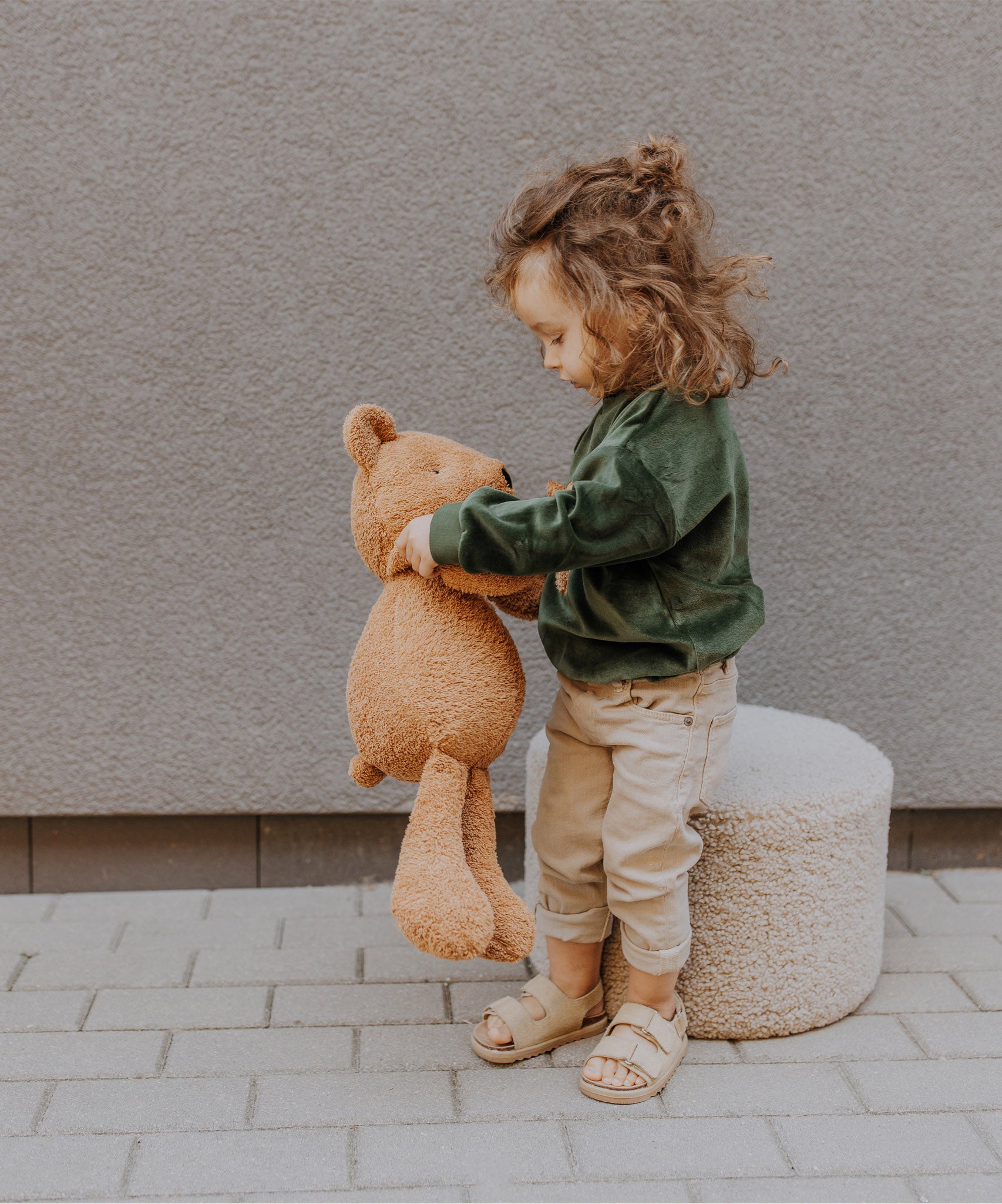 A child standing on a stone surface and playing with the Wooly Organic soft teddy bear in caramel available at Babipur