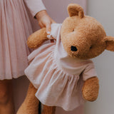 A close up of the Wooly Organic soft teddy bear in caramel brown with a pink tutu available at Babipur showing a child holding by its hand and cute facial features.