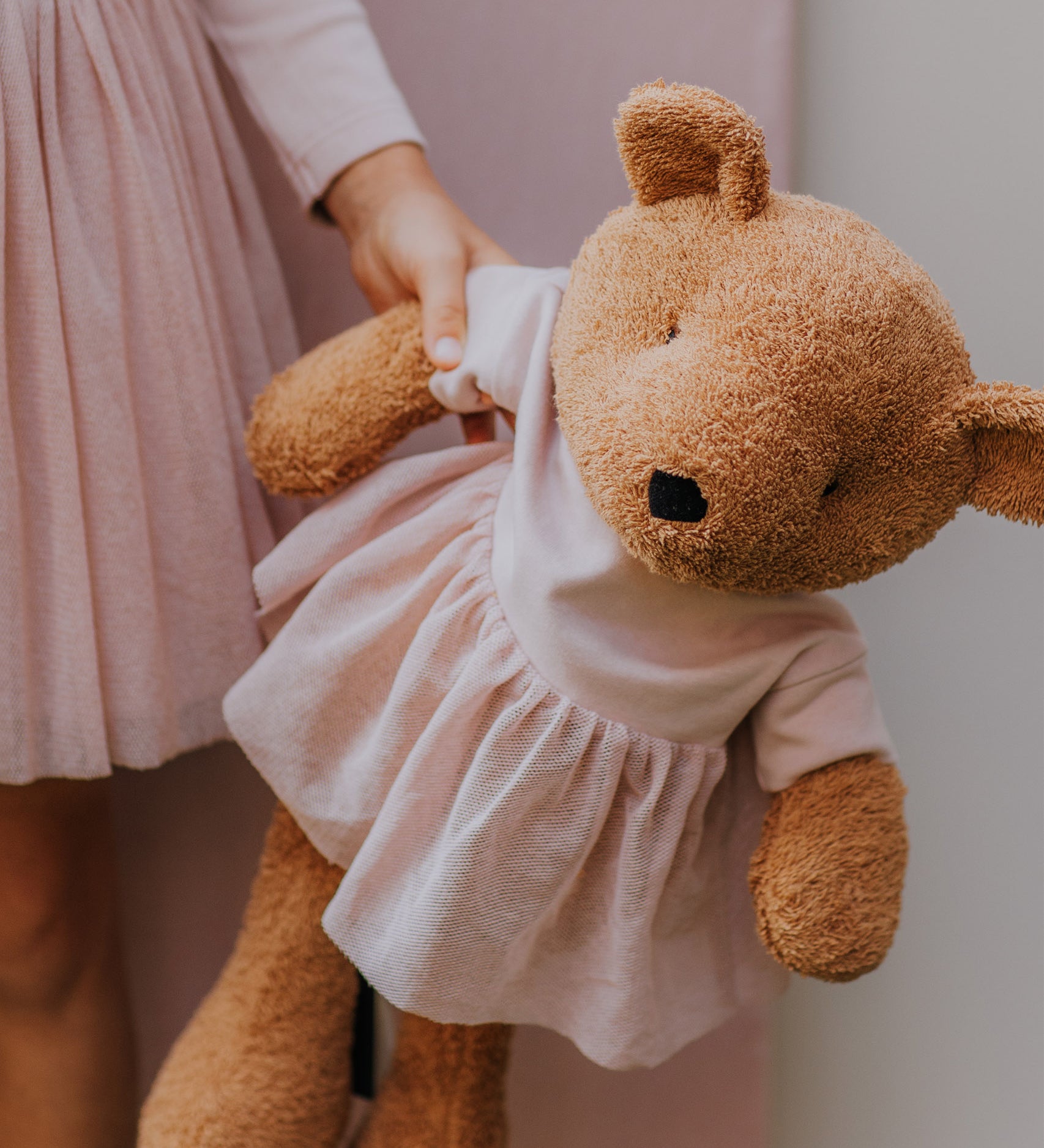 A close up of the Wooly Organic soft teddy bear in caramel brown with a pink tutu available at Babipur showing a child holding by its hand and cute facial features.