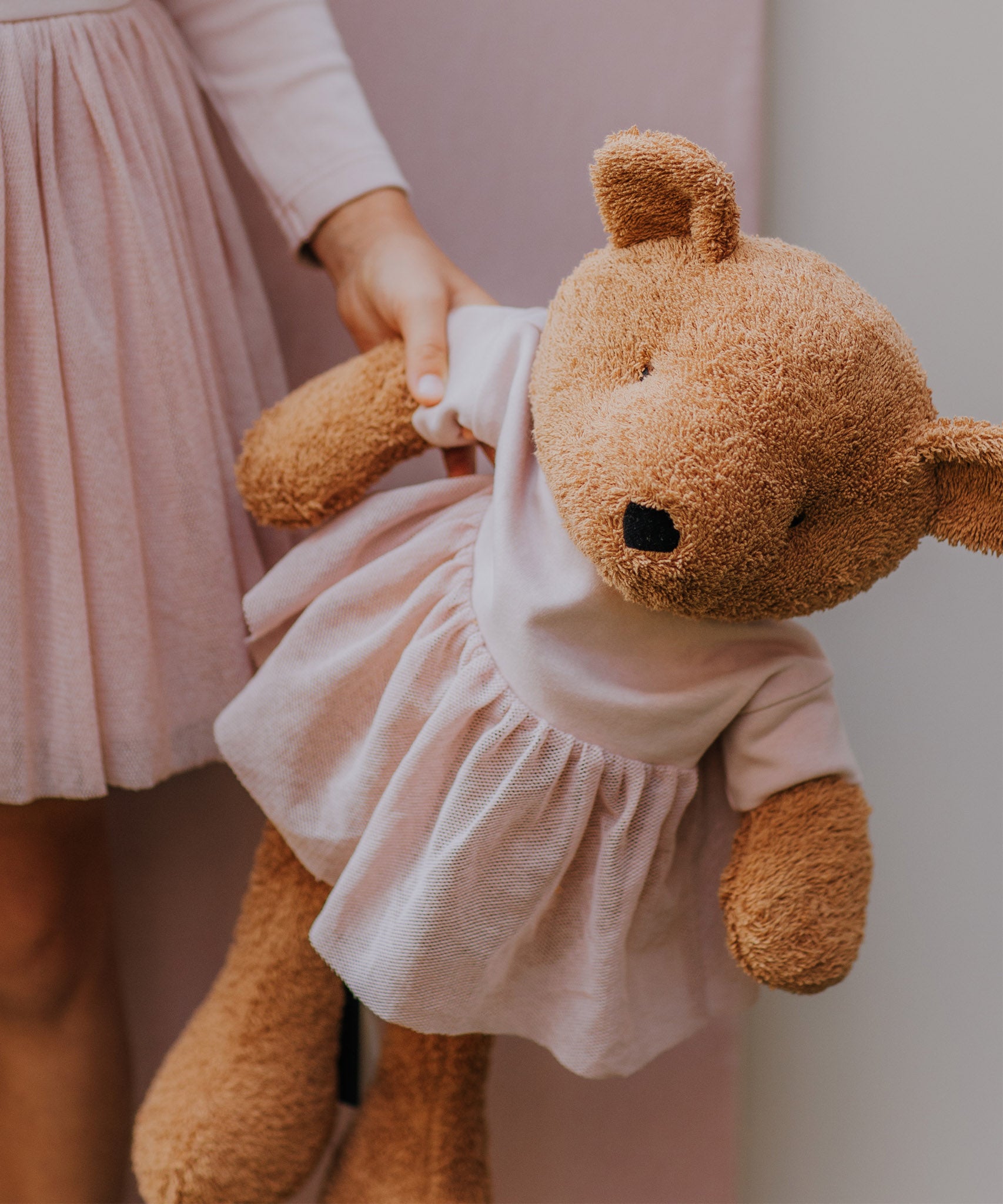 A close up of the Wooly Organic soft teddy bear in caramel brown with a pink tutu available at Babipur showing a child holding by its hand and cute facial features.