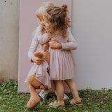 2 children hugging and holding the Wooly Organic soft teddy bear in caramel brown with a pink tutu available at Babipur. 