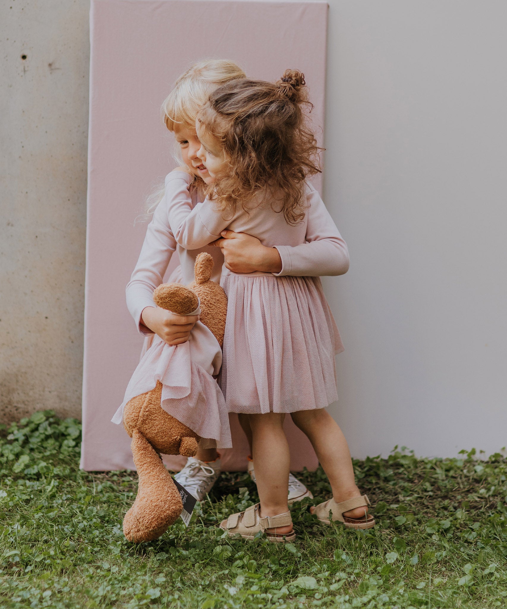 2 children hugging and holding the Wooly Organic soft teddy bear in caramel brown with a pink tutu available at Babipur. 