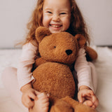 A close up of a child smiling and hugging the Wooly Organic soft teddy bear in caramel brown available at Babipur.