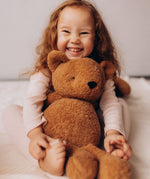 A close up of a child smiling and hugging the Wooly Organic soft teddy bear in caramel brown available at Babipur.