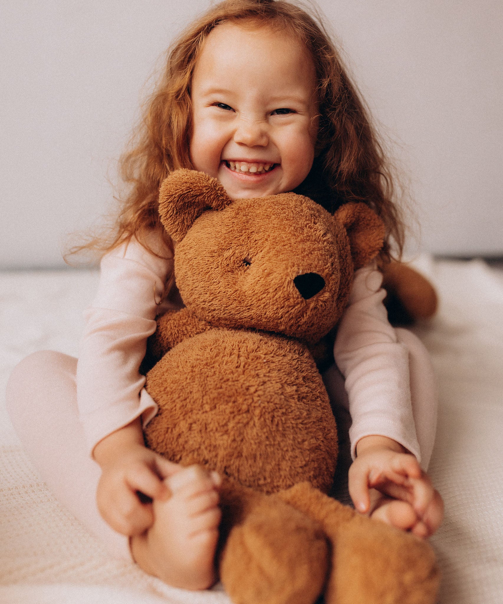 A close up of a child smiling and hugging the Wooly Organic soft teddy bear in caramel brown available at Babipur.