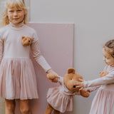 2 children holding the Wooly Organic soft teddy bear in caramel brown with a pink tutu by the hand. Teddy bear available at Babipur. 