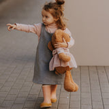A small child walking down the street and holding the Wooly Organic soft teddy bear in caramel brown with a pink tutu available at Babipur. 
