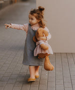 A small child walking down the street and holding the Wooly Organic soft teddy bear in caramel brown with a pink tutu available at Babipur. 
