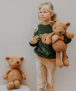 A child wearing the Wooly Organic tea leaf velour embroidered sweater and holding the soft teddy bear in caramel, both available at Babipur. 