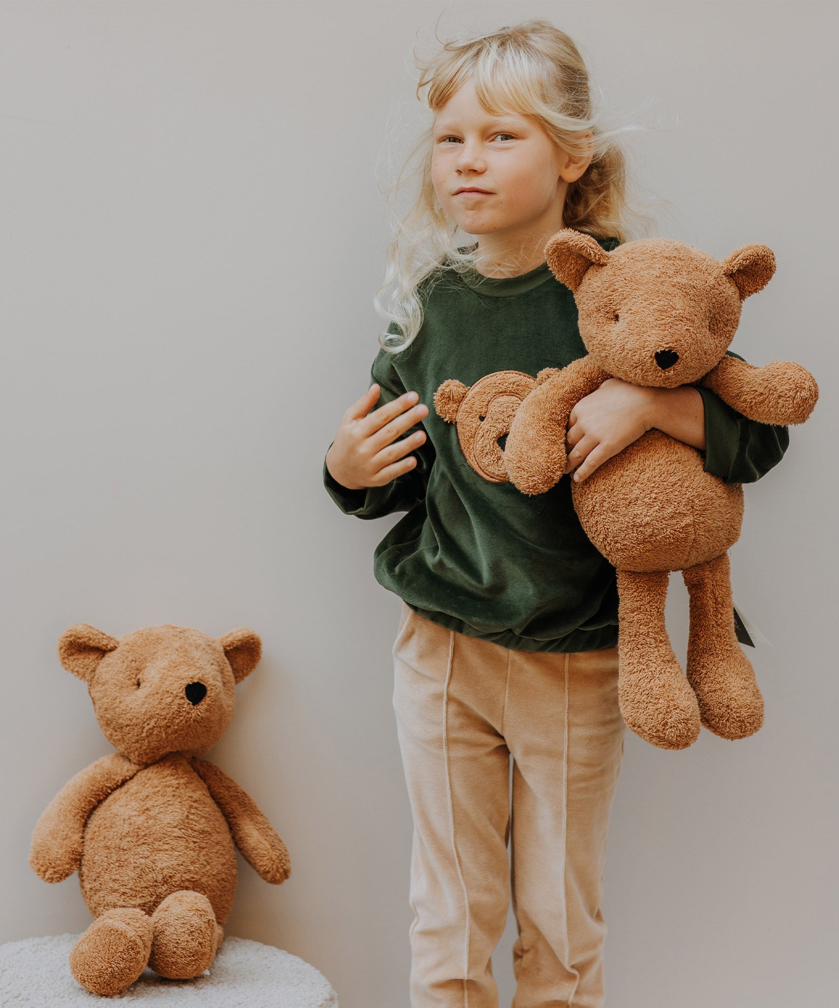 A child wearing the Wooly Organic tea leaf velour embroidered sweater and holding the soft teddy bear in caramel, both available at Babipur. 