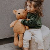 A child sitting on a soft cushion and holding the soft Wooly Organic teddy bear in caramel available at Babipur