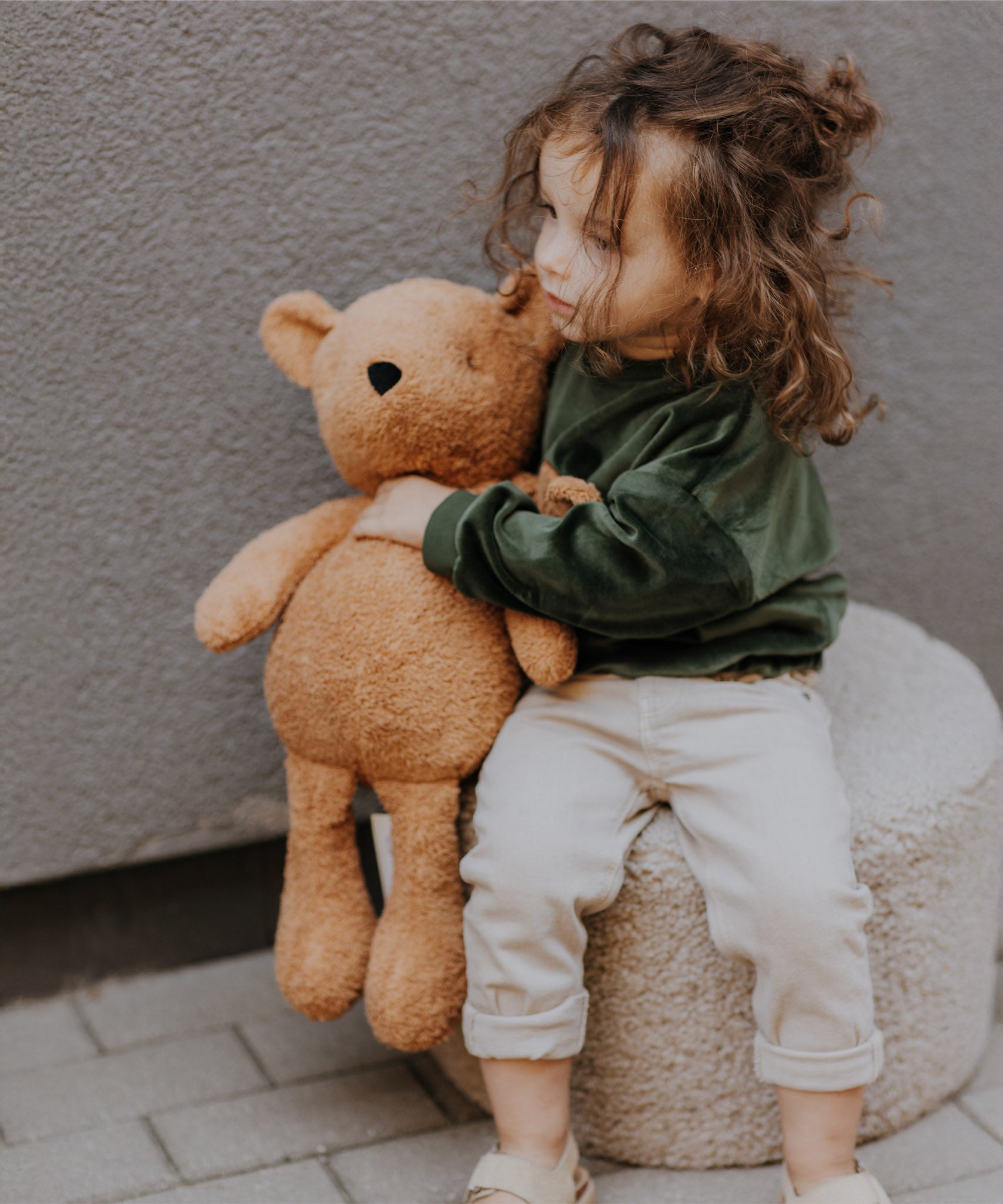 A child sitting on a soft cushion and holding the soft Wooly Organic teddy bear in caramel available at Babipur