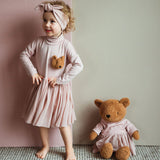 A child posing next to the Wooly Organic soft teddy bear in caramel brown with a pink tutu sitting down. Teddy bear available at Babipur. 