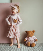 A child posing next to the Wooly Organic soft teddy bear in caramel brown with a pink tutu sitting down. Teddy bear available at Babipur. 