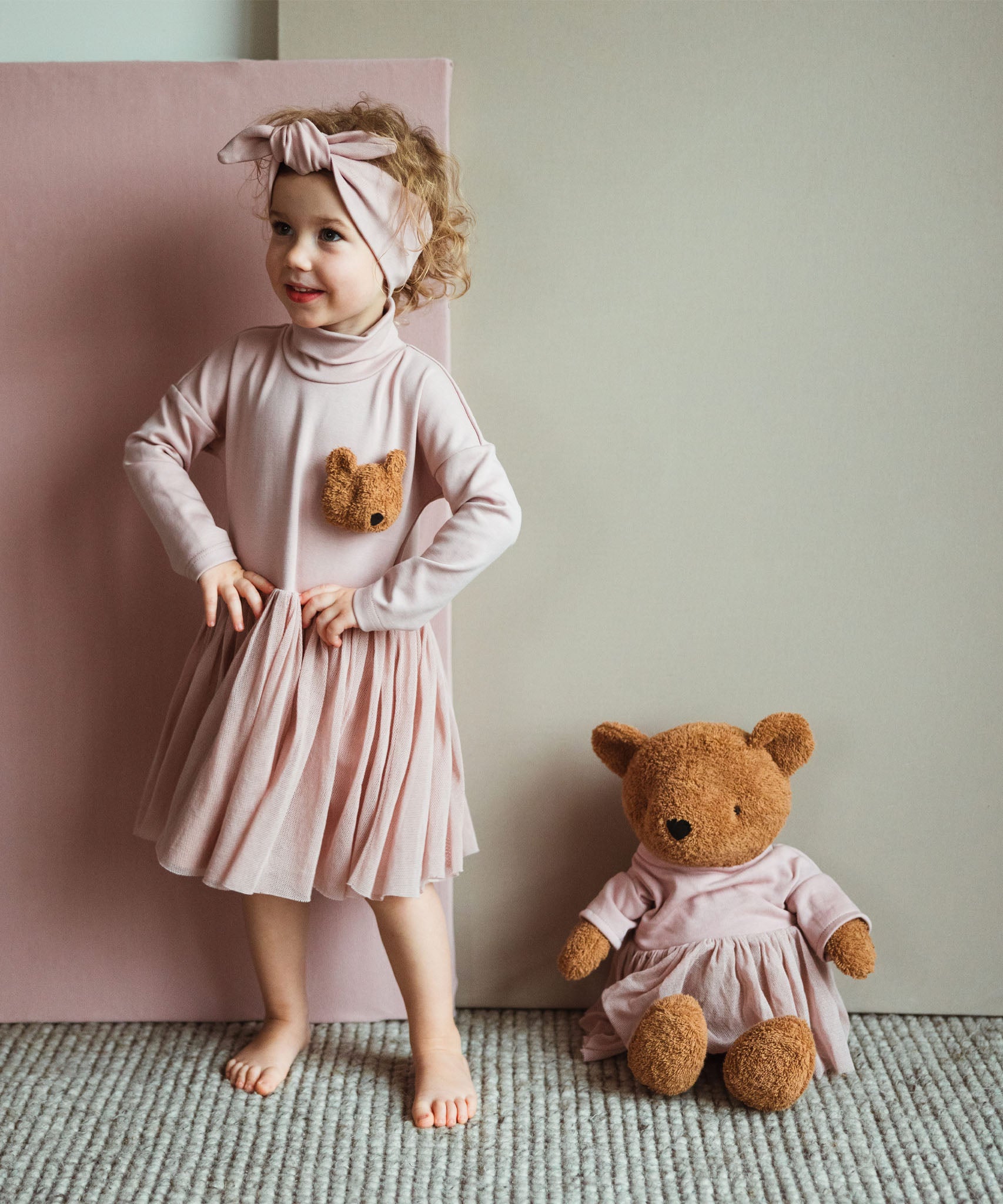 A child posing next to the Wooly Organic soft teddy bear in caramel brown with a pink tutu sitting down. Teddy bear available at Babipur. 