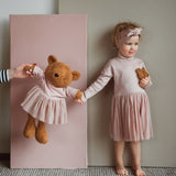 A child standing next to the Wooly Organic soft teddy bear in caramel brown with a pink tutu and holding by the hand. Teddy bear available at Babipur. 